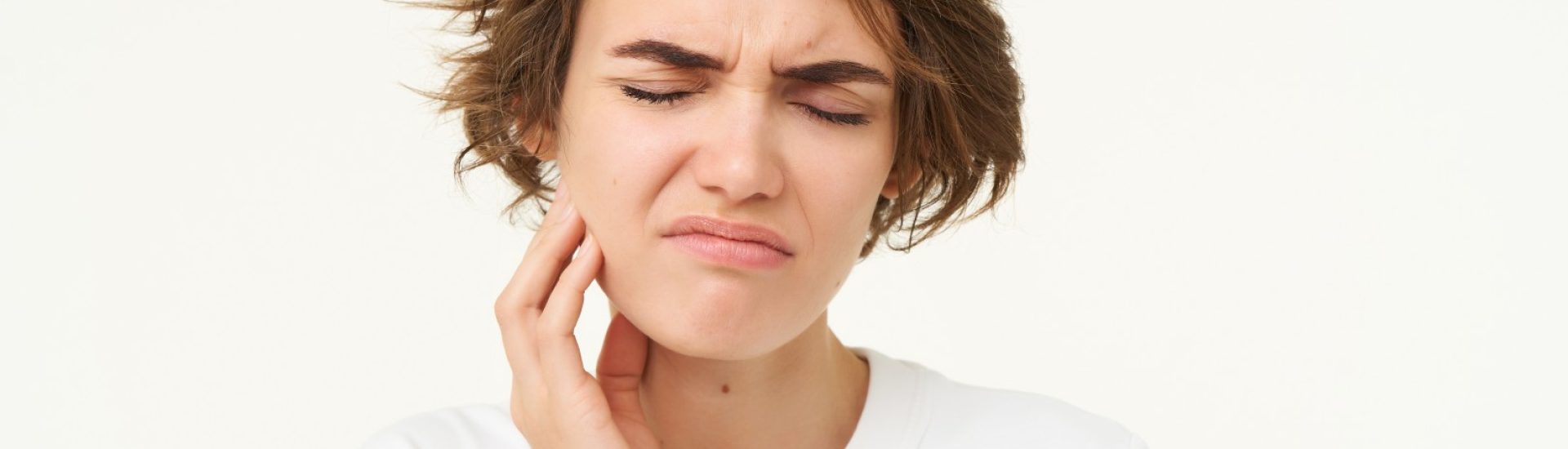 Close up of woman has a toothache, touches her teeth and frowns from painful discomfort, stands over white background.
